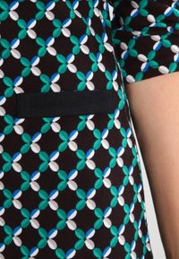 Korte mouwen top met een geometrisch patroon in groen, blauw en wit op een donkerbruine achtergrond; inclusief een zwart zakdetail.