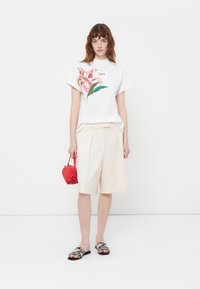 T-shirt bianca a maniche corte con ricamo floreale, pantaloni beige su misura, ciabatte a righe in bianco e nero e una piccola borsa rossa.