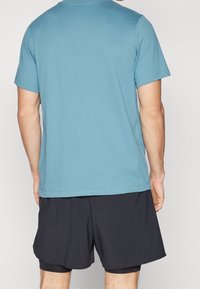 Muž v světlomodrém tričku s krátkým rukávem a černých sportovních šortkách, zobrazený zezadu na jednolitém pozadí.