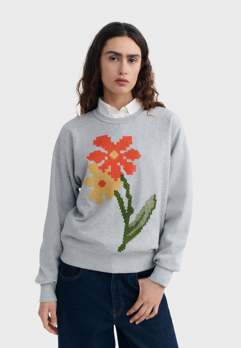 Femme portant un pull gris clair orné de fleurs pixelisées rouges et jaunes, une chemise blanche à col, et un jean bleu foncé, debout devant un fond uni.