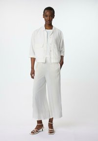 Veste blanche à manches courtes avec deux poches, associée à un pantalon clair à jambes larges et des sandales à lanières. Texture de tissu lisse.