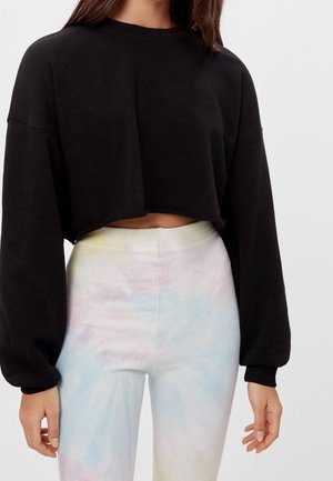 Femme portant un pull court noir à manches longues et un pantalon taille haute tie-dye pastel sur un fond uni.