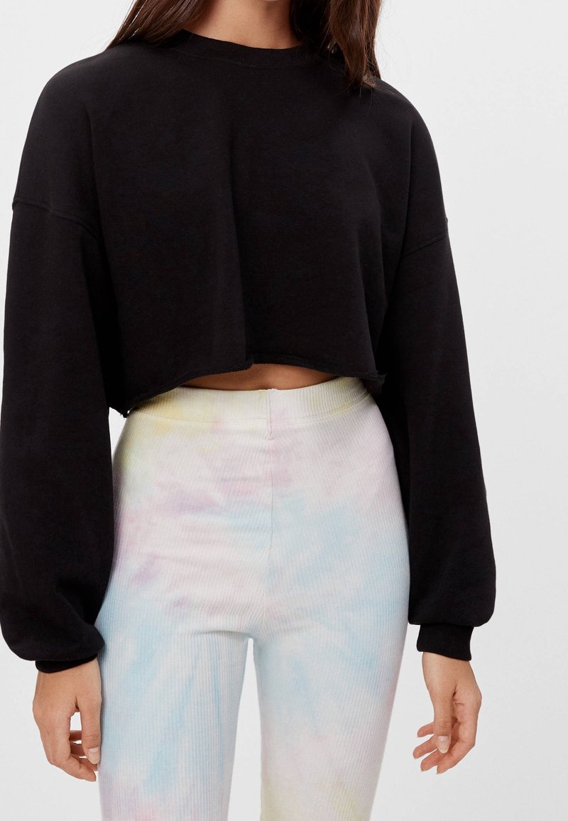 Femme portant un pull court noir à manches longues et un pantalon taille haute tie-dye pastel sur un fond uni.