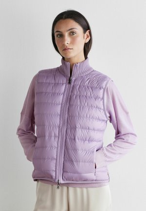 Frau trägt eine hellviolette, wattierte ärmellose Weste mit Reißverschluss, steht mit den Händen in den Taschen vor einem schlichten weißen Hintergrund.