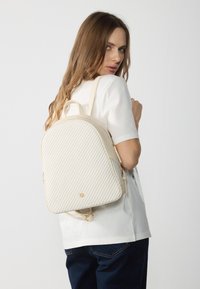 Béžový batoh s texturovanými diagonálními pruhy, zaobleným tvarem, zlatými kovovými detaily a nastavitelnými popruhy, nošený na zádech modelky.