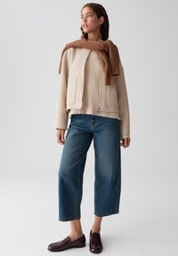 Donna che indossa una giacca beige con zip sopra un maglione abbinato, sciarpa marrone, jeans cropped blu a gamba larga e mocassini marroni scuri, in posizione eretta.