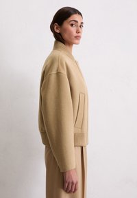 Beige Wollmischung Bomberjacke mit ribbeltem Kragen, seitlichem Taschen und lässiger Passform, auf einem Modell vor einem hellen Hintergrund präsentiert.