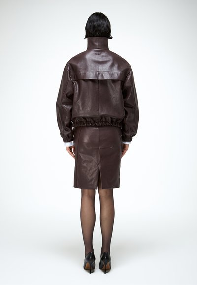 Rövid fekete hajú személy sötétbarna bőrdzsekit és hozzá illő szoknyát visel, fekete átlátszó harisnyával és fekete magassarkú cipővel, háttal áll.