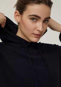 Felpa blu navy in morbido tessuto, con tasca a marsupio e cappuccio regolabile con cordino. Inquadratura ravvicinata del viso con trucco naturale.