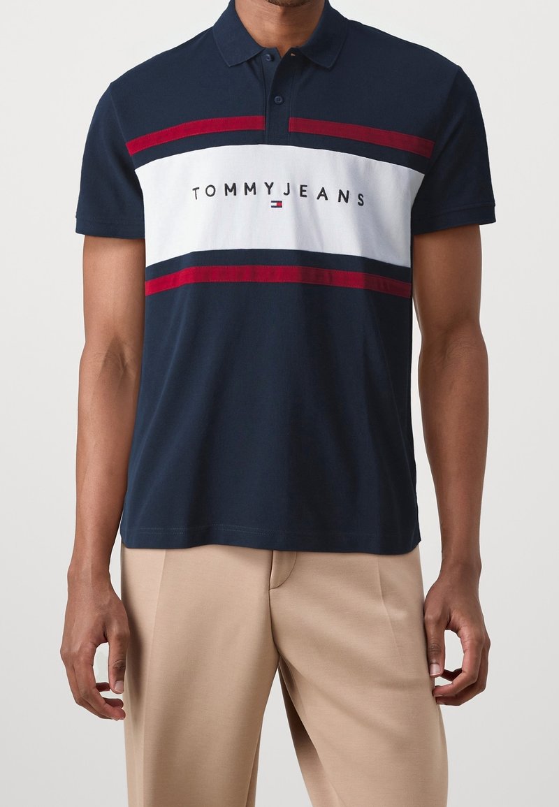 Polo azul marino de mezcla de algodón con rayas horizontales blancas y rojas. Cuenta con cuello, mangas cortas y el logo 'TOMMY JEANS' en el pecho.