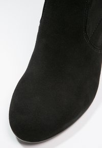 Primer plano de una bota negra de ante hasta el tobillo que muestra la puntera redondeada y los detalles de costura en la parte superior.