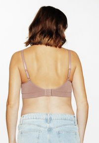 Donna con capelli castani lunghezza spalle, che indossa un reggiseno color mauve e jeans azzurri chiari, vista da dietro su uno sfondo bianco.