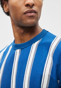 Camisola azul com gola redonda, riscas verticais brancas e finos detalhes em azul-petróleo. Textura em malha com gola canelada, apresentando um corte ajustado.