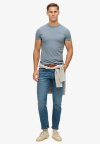 T-shirt gris à manches courtes, jeans bleus, pull gris clair noué autour de la taille et baskets beiges. Tenue simple et décontractée.