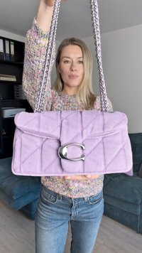 Lila prošívaná látková kabelka se stříbrným sponovým zapínáním ve tvaru C a řetízkovým popruhem, má obdélníkový tvar a hladkou strukturu.