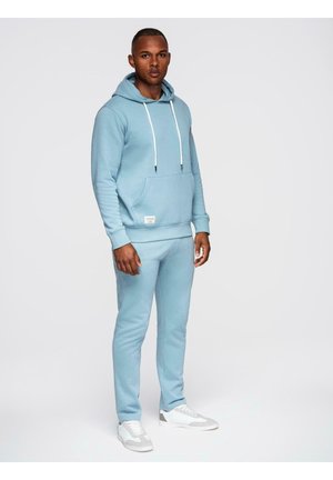 Mand stående iført lyseblå hættetrøje og matchende joggingbukser med hvide sneakers på ensfarvet baggrund.