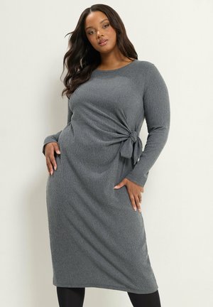 Femme aux longs cheveux foncés portant une robe en maille côtelée grise à manches longues avec un détail noué sur le côté, posant devant un fond clair uni.