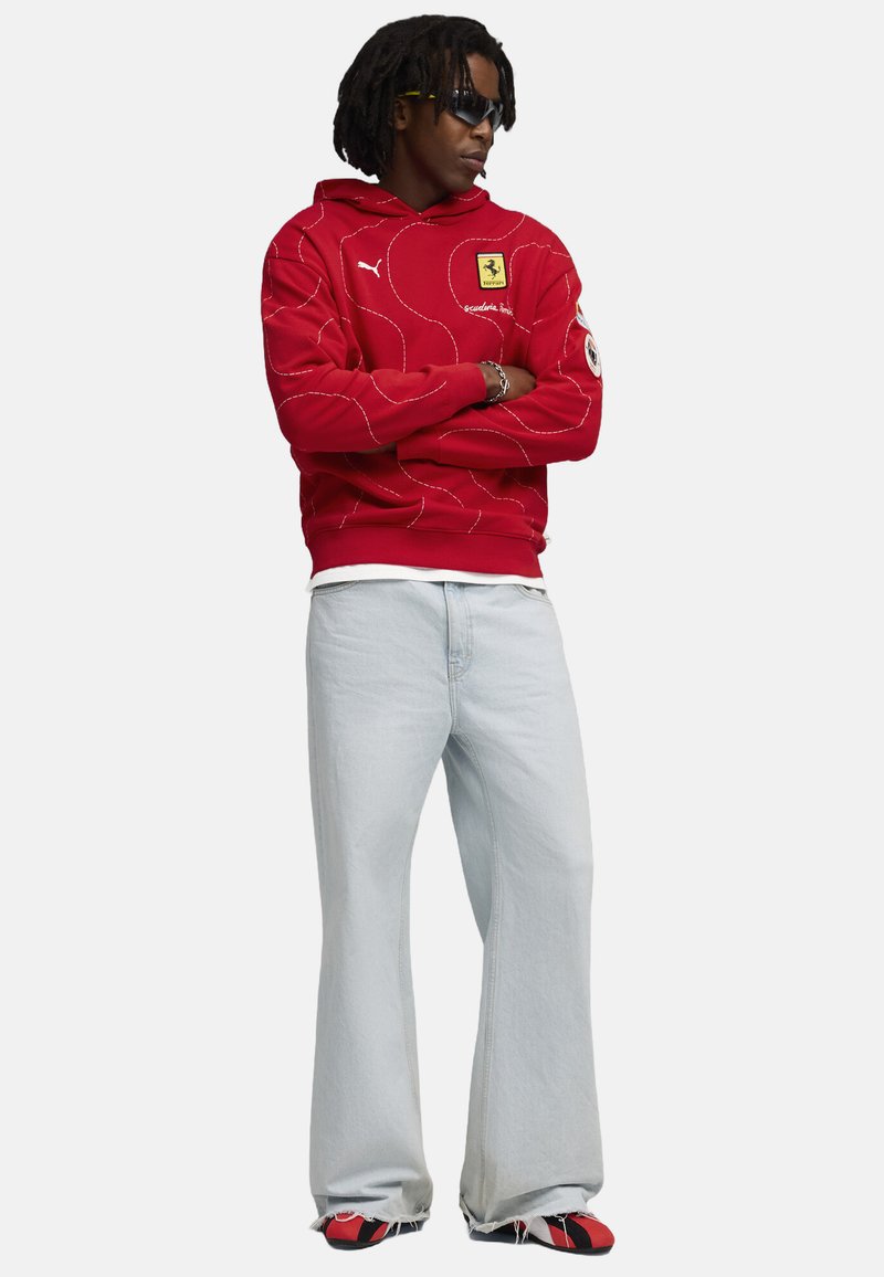 Rode hoodie met witte stiksels en Ferrari-logo, gecombineerd met lichtblauwe flared jeans en rood-zwart sneakers. Zwarte zonnebril maakt de look compleet.