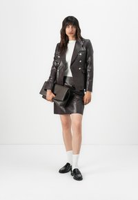 Tmavohnedý kožený blazer s textúrovaným povrchom, skombinovaný s matching koženou sukňou. Model drží sivú kabelku, má na sebe biele ponožky a čierne topánky.