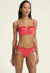 Jasnoróżowy bikini z falbankami. Górna część ma usztywniony fason z cienkimi ramiączkami, a dół ma wiązania po bokach z dopasowanymi falbankami.