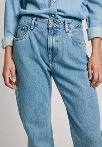 Gai zila denim auduma bikses ar augstu jostas vietu, piecu kabatu dizainu un nelielu nolietojumu. Iezīmēts ar vara toņu detaļām un klasiskām piedurknēm.