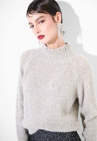Maglione a maglia grigio chiaro con textura, collo alto, con fili metallici e vestibilità comoda, abbinato a orecchini pendenti intricati.