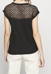 Mustat lyhyt hihaiset paita, jossa on läpinäkyvä pitsi takaosa, jossa on sahalaitakuviota, yhdistettynä vaaleanvärisiin corduroy-housuihin.