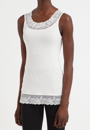 Biały top bez rękawów z koronkową lamówką na dekolcie i krawędzi, noszony przez osobę o ciemnej karnacji, ubrana w czarne spodnie, stojącą na jednolitym tle.