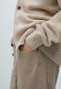 Beżowy sweter na guziki z prążkowanymi mankietami oraz ręką w kieszeni jasnobrązowych spodni z tkaniny. Gładka tekstura i swobodny krój.