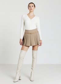 Weiße Strickjacke mit V-Ausschnitt, kombiniert mit einem beigen, plissierten Rock mit Taillengürtel und weißen überknielangen, hochhackigen Stiefeln.