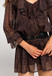 Vestido marrón con estampado, con un escote con volantes y mangas largas transparentes, ceñido en la cintura por un cinturón de cuero negro con herrajes dorados.
