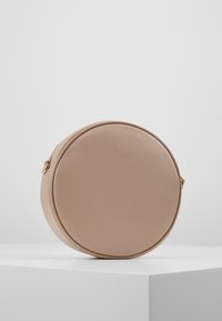 Runde, beige Umhängetasche aus glattem, synthetischem Material mit einem goldenen Reißverschluss und Akzenten. Minimalistisches Design, kompakte Form.