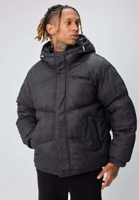 Veste matelassée noire avec un motif en losange texturé, dotée d'une capuche, d'une fermeture éclair et de deux poches avant. Le matériau semble doux et isolant.