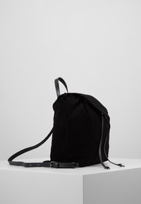 Černý semišový batoh s koženými popruhy a sponou, stojící vzpřímeně na bílém podstavci na jednoduchém světle šedém pozadí.