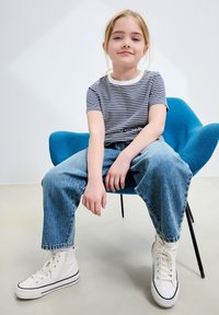 Koszulka w paski z krótkim rękawem i białym kołnierzykiem, jasnoniebieskie jeansy z wysokim stanem oraz białe trampki za kostkę na niebieskim krześle.