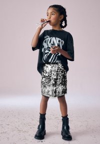 Schwarzes übergroßes Rolling Stones T-Shirt, silberner Pailletten-Minirock, schwarze Stiefeletten, geschichtete Socken, stehend mit einem Partybläser.