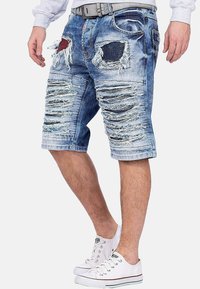 Shorts en denim présentant des détails usés, des bords effilochés et plusieurs poches, de couleur bleu clair avec une coupe décontractée. Associés à des baskets blanches.