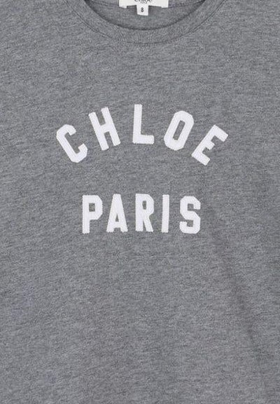 Szary materiał z białymi, dużymi literami pisanymi jako "CHLOE", wygięty nad "PARIS" w prostej linii na odzieży.