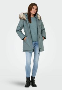 Cappotto lungo imbottito in un verde teal smorzato con cappuccio in finta pelliccia. Abbinato a un maglione blu, jeans chiari e stivali neri alla caviglia.