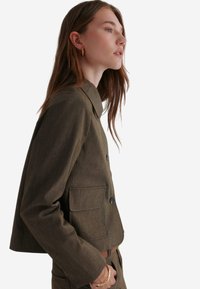 Olivgrüne, kurz geschnittene Jacke mit Kragen, zwei vorderen Taschen und Knopfverschluss. Glattes Textilmaterial, figurbetontes Design.