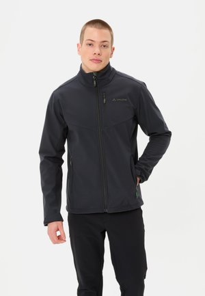 Giovane uomo in piedi con una mano in tasca, indossa una giacca con zip grigio scuro e pantaloni neri su uno sfondo bianco semplice.