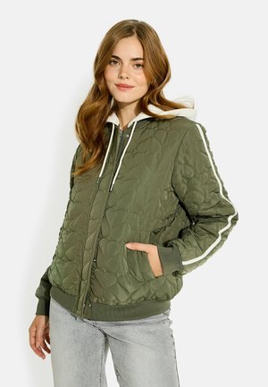 Joven mujer con chaqueta acolchada verde oliva con estampados de corazones y sudadera con capucha blanca, de pie con una mano en el bolsillo.