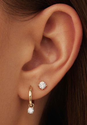 Boucle d'oreille en or avec un diamant pendant, à côté d'une boucle d'oreille puce avec un diamant rond, tous deux montés sur quatre griffes.