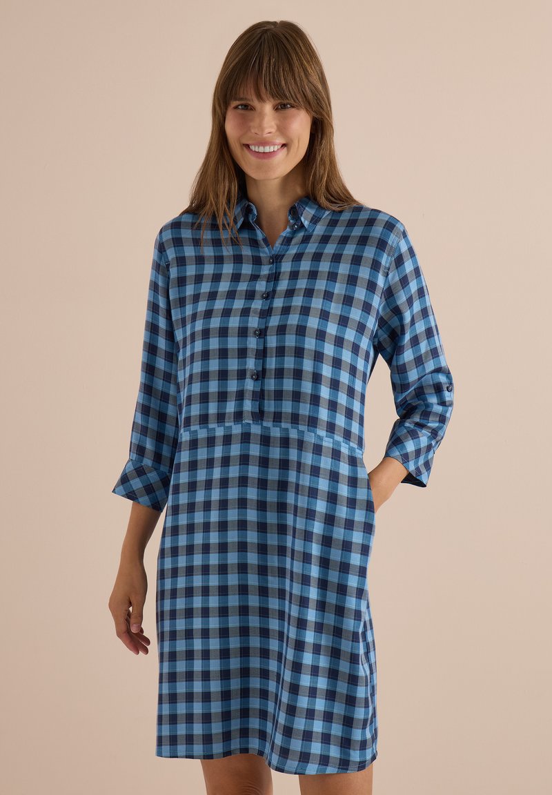 Femme souriante portant une robe-chemise à manches longues à carreaux bleus et noirs avec des boutons, debout devant un fond beige uni.