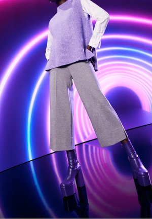 Un gilet tricoté violet clair superposé sur une chemise blanche, associé à un pantalon large gris et des bottes plateformes violettes brillantes sur fond néon vibrants.