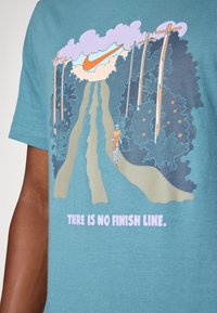 Camiseta de algodón en color agua con un gráfico de un corredor en un sendero rodeado de árboles, nubes y la frase "NO HAY LÍMITE DE META" en color rosa.