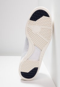 Suela de zapatilla blanca con goma texturizada y patrón hexagonal. Presenta un acento azul oscuro en el talón y una entresuela blanca lisa.