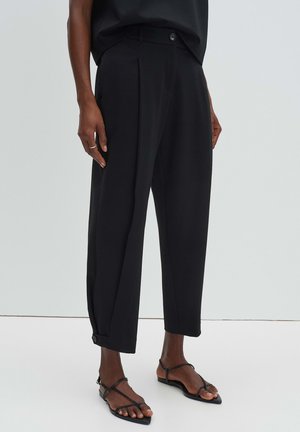 Personne portant un pantalon noir raccourci avec des poignets boutonnés et des sandales plates noires à brides, debout devant un fond gris uni.