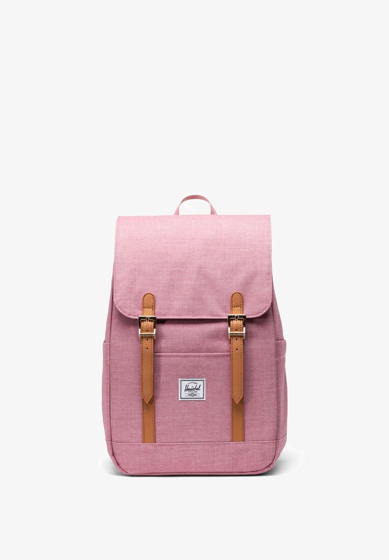 Różowy plecak materiałowy z dwoma brązowymi skórzanymi paskami i metalowymi klamrami. Posiada górną klapę oraz przednią kieszeń z naszywką z logo.