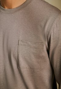 Camicia grigia in cotone con scollo rotondo, dotata di una tasca frontale e una finitura morbida e testurizzata. Design semplice, adatto per un abbigliamento casual.